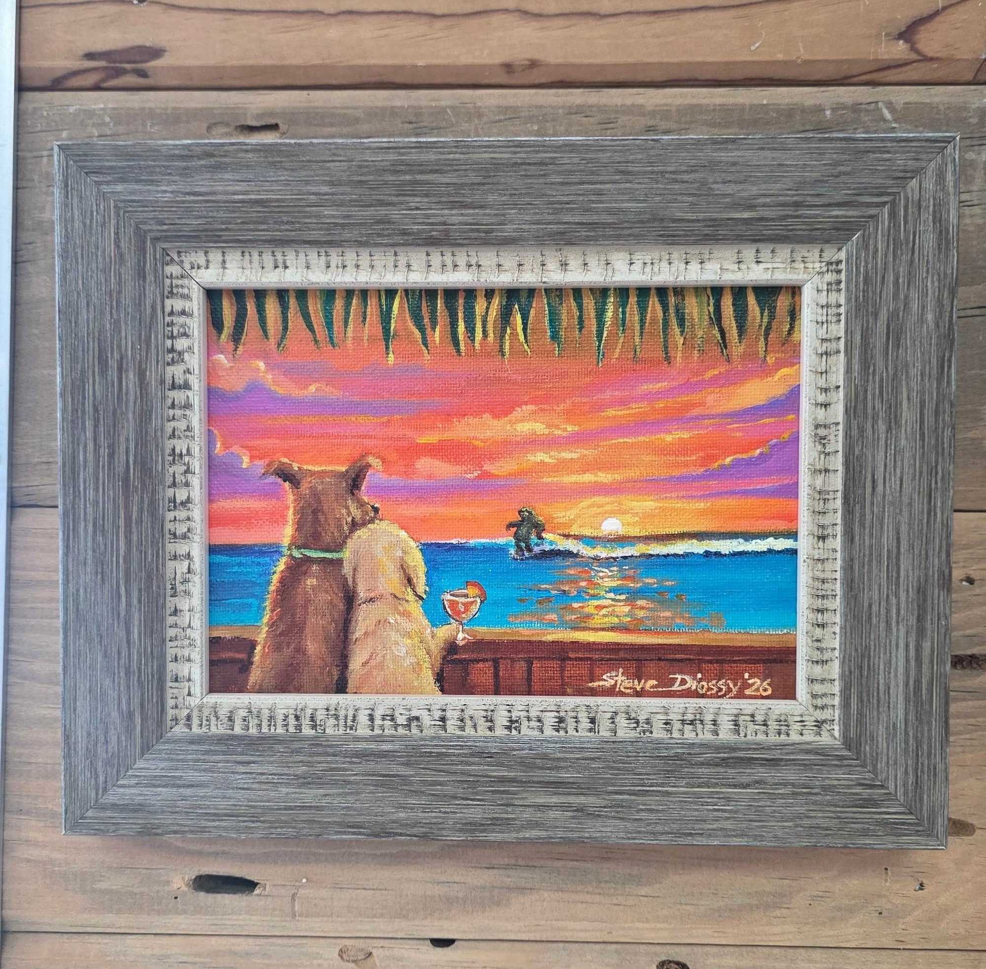 A framed art print depicting a beach sunset scene with two dogs looking at the ocean while a sloth surfs in the waves. Hung on a rustic wooden plank wall.